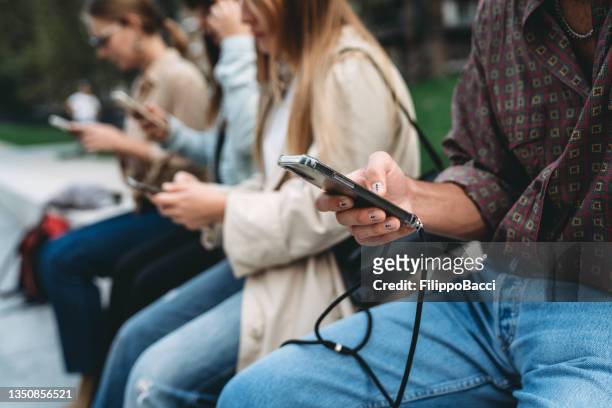 vier freunde benutzen smartphones, während sie in einem öffentlichen park in der stadt sitzen - propaganda stock-fotos und bilder
