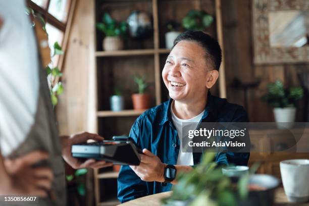 cheerful senior asian man paying bill with credit card contactless payment on smartphone in a cafe, scanning on a card machine. electronic payment. elderly and technology - scanner stock stock pictures, royalty-free photos & images