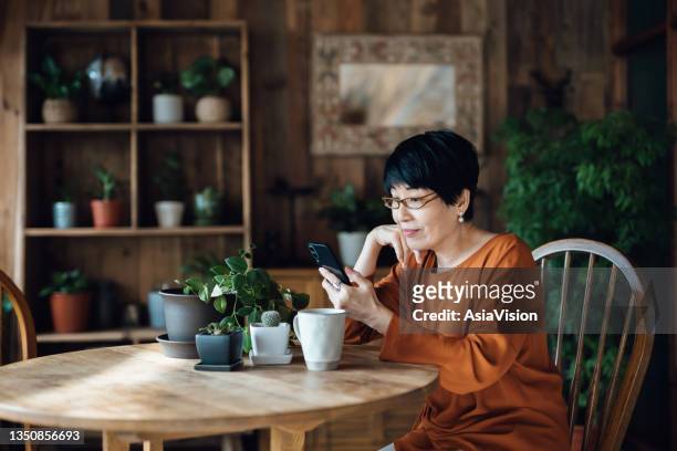 smiling senior asian woman sitting at the table, surfing on the net and shopping online on smartphone at home. elderly and technology - asian woman stock pictures, royalty-free photos & images