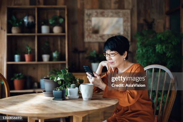 mulher asiática sênior sorridente sentada à mesa, surfando na internet e fazendo compras online no smartphone em casa. idosos e tecnologia - reading glasses - fotografias e filmes do acervo