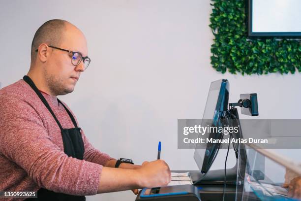 joven barista de pie detrás de la caja registradora atendiendo al cliente y haciendo el pedido en el ordenador - ordenador stock-fotos und bilder