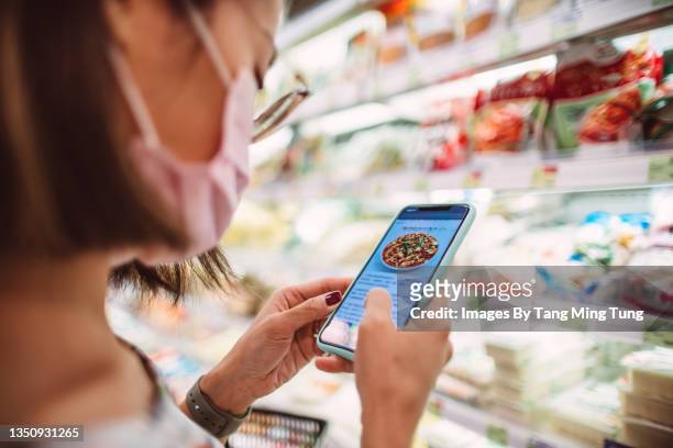 young woman in protective face mask checking recipe on her smartphone and shopping for ingredients in supermarket - レシピ ストックフォトと画像