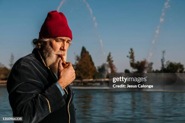 old man, with a big and large beard, dressed as a sailor, with a smoking pipe. with a lake in the background. old men, beard, bearded, hipster, barber and retirement concept. copyspace - big beard stockfoto's en -beelden