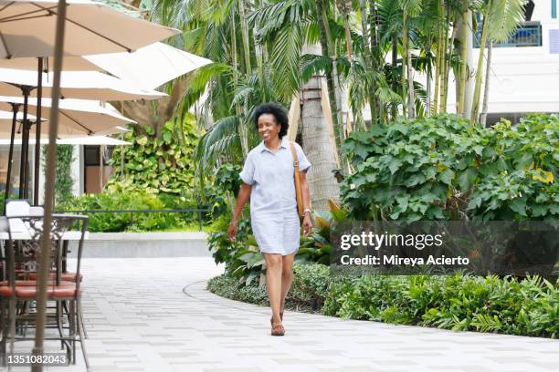 an african american mature woman with curly hair, walks through a commercial area while wearing a light blue striped dress and leather purse. - west palm beach florida stock pictures, royalty-free photos & images