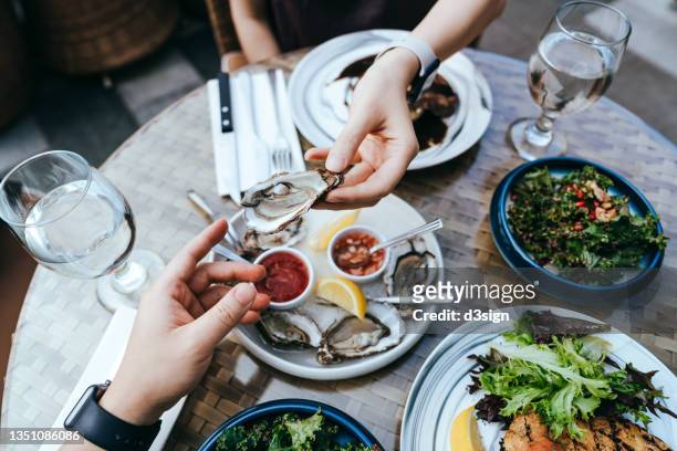 high angle view of woman passing fresh oyster to man across the dining table during lunch, enjoying a scrumptious meal in outdoor restaurant. sharing and togetherness. eating out lifestyle. outdoor dining concept - premium dining stockfoto's en -beelden
