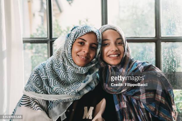 portrait of two sisters with hijabs against a big window - moroccan culture stock pictures, royalty-free photos & images