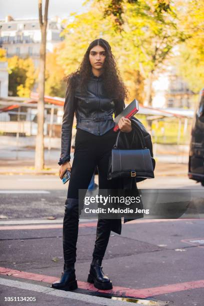 Model Anita Pozzo wears a black leather belted jacket, black pants, thigh-high black leather boots, black Balenciaga Hourglass bag, and carries...