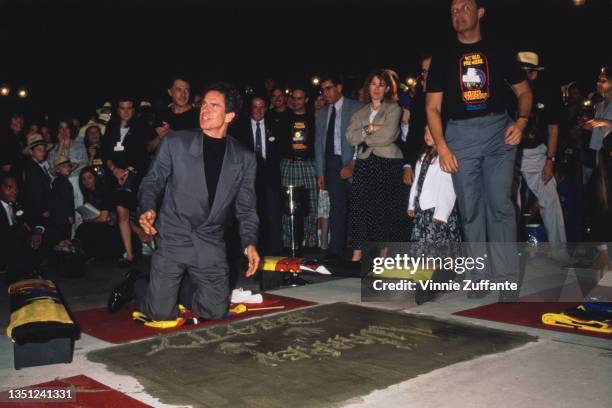 American actor Warren Beatty, wearing a grey suit over a black crew neck sweater, kneeling on the ground having signed his name in cement and left...