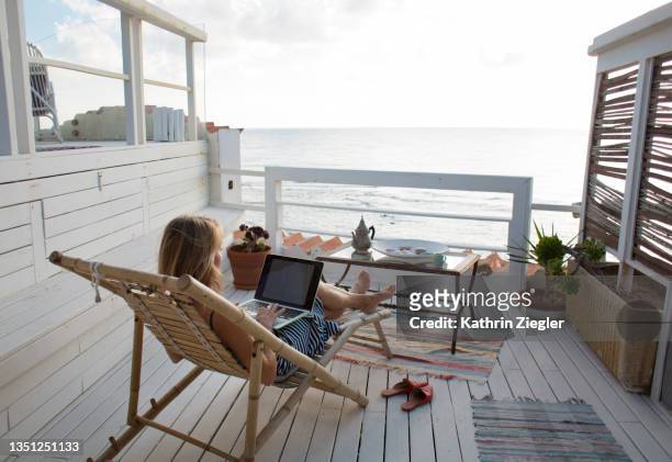 woman sitting on deck chair on a terrace with sea view, using laptop computer - isolamento foto e immagini stock