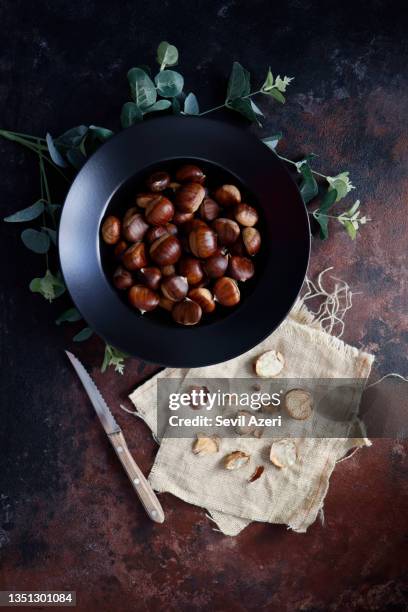 chestnuts in a black deep plate on a rough concrete floor with a black-brown rust effect. next to the plate are peeled chestnuts and their shells on burlap with a wooden-handled knife. - chestnut stock pictures, royalty-free photos & images