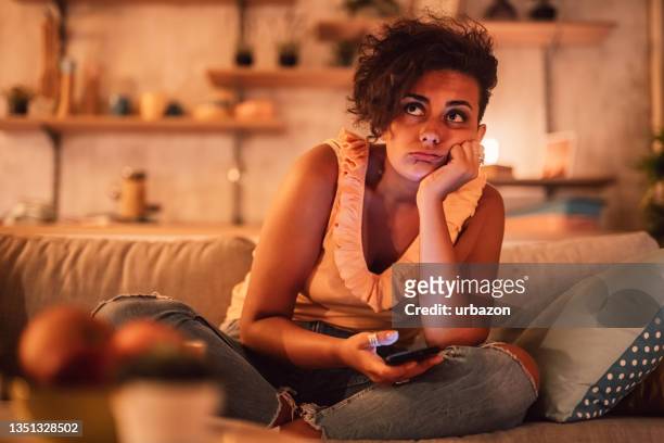 bored woman sitting on sofa and holding phone - bored imagens e fotografias de stock