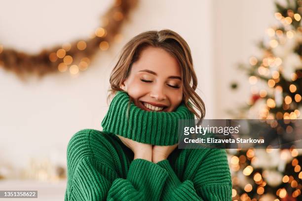 portrait of a funny emotional cheerful young woman with horns on her head in a green cozy warm sweater laughing having fun holding and eating a lollipop and ginger cookies on christmas day on the background of a decorated christmas tree at home in winter - before christmas foto e immagini stock