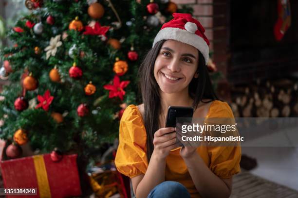beautiful thoughtful young woman looking up smiling sitting on the floor next to the christmas tree and presents, wearing a santa's hat while holding her smartphone - chat noel stockfoto's en -beelden