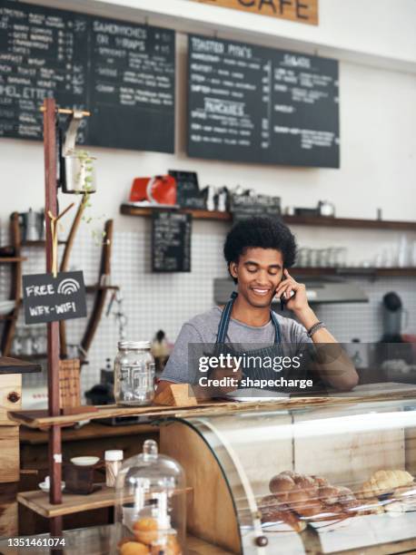 shot of a young male waiter using a notebook while on a call at a cafe - restaurant manager phone stock pictures, royalty-free photos & images