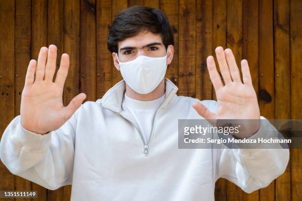 young latino man wearing a face mask, looking at the camera, and gesturing to stop - avoiding danger stock pictures, royalty-free photos & images