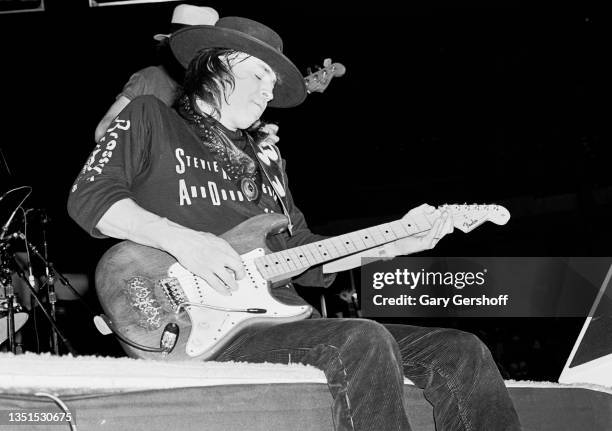 Sitting onstage, American Blues and Rock musician Stevie Ray Vaughan plays guitar with his band Double Trouble during the 'Texas Flood' tour at Byrne...