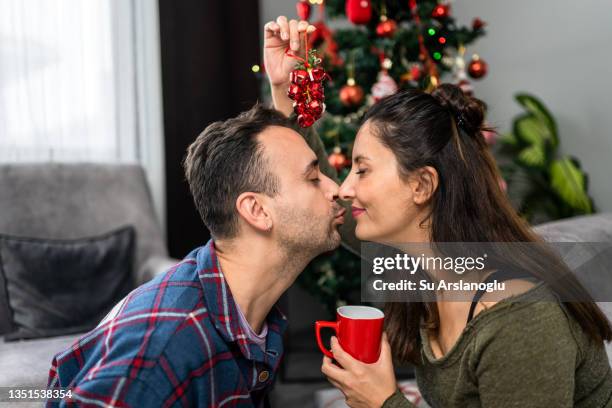 young couple decorating christmas tree at home and drinking coffee - mistletoe kiss stockfoto's en -beelden