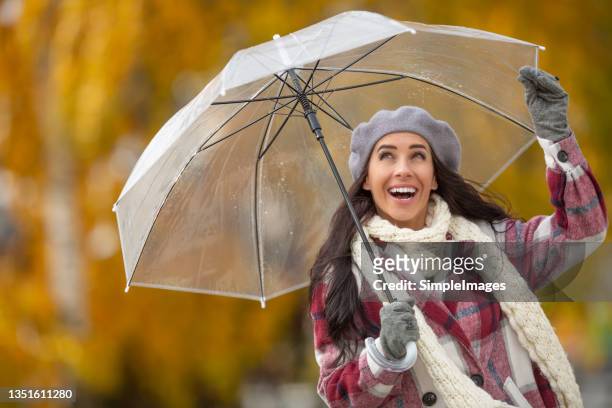 happy woman smiles and looks up at open umbrella above her sourrounded by colorful autumn trees. - female fashion with umbrella stock pictures, royalty-free photos & images