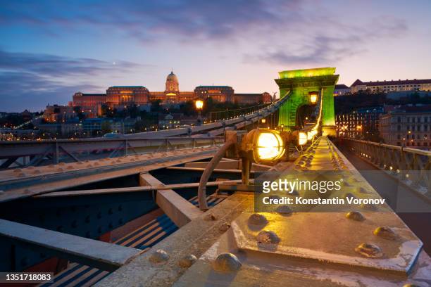 night illumination on the chain bridge in budapest. lamps and construction elements in the foreground.  royal palace on castle hill on the background - hungarian culture stock-fotos und bilder