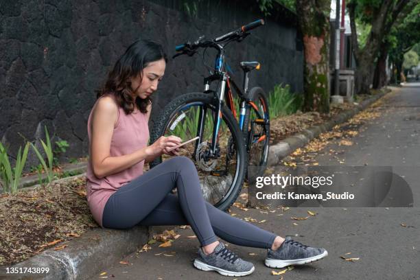 millenial asian woman sitting on the  curb using phone for trading - java oeste imagens e fotografias de stock