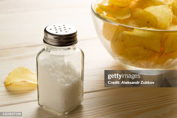 crispy golden fried fat potato chips in a glass bowl or plate, on a white wooden background or table. next to the salt shaker. the concept of unhealthy diet and lifestyle, the accumulation of excess weight. - salt shaker stockfoto's en -beelden