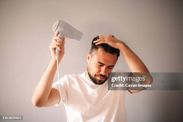 man blow drying hair against gray wall at home - föhn stock-fotos und bilder