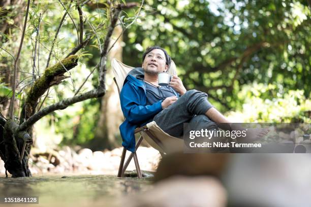 man relaxing sitting on a camping chair while drinking coffee and looking a view in a morning at a camping site in the forest. natural therapy and mental health recovery. - アウトドア　日本人 ストックフォトと画像