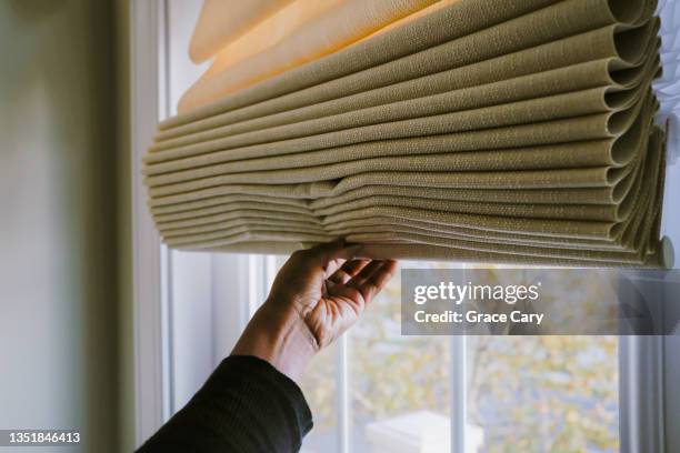 woman adjusts roman shade on window - curtain foto e immagini stock