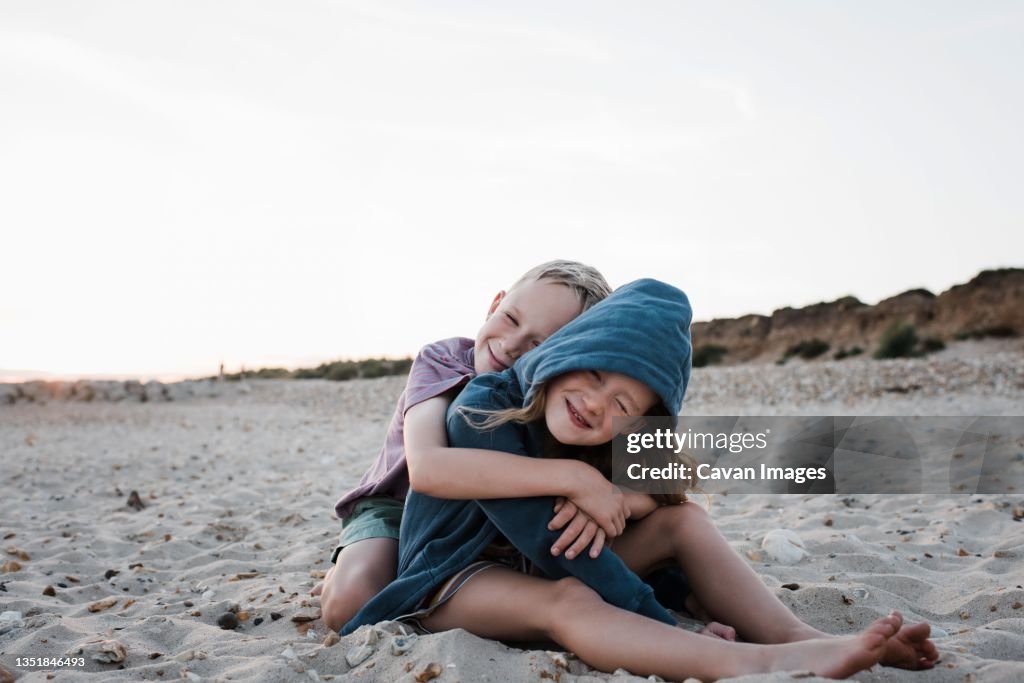 Brother and sister sat on sand playfully hugging at the beach