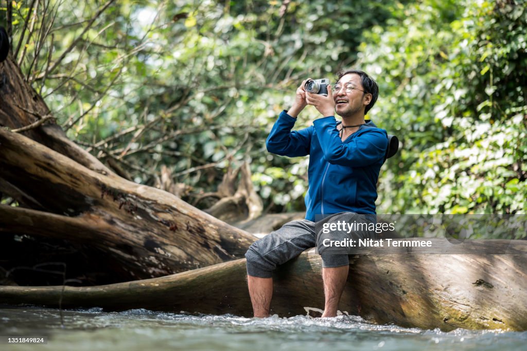 Senior Backpacker enjoying nature while taking photos with a mirrorless camera in a rainforest during camping. Camping Lover, Nature Addicted, and Simply living.
