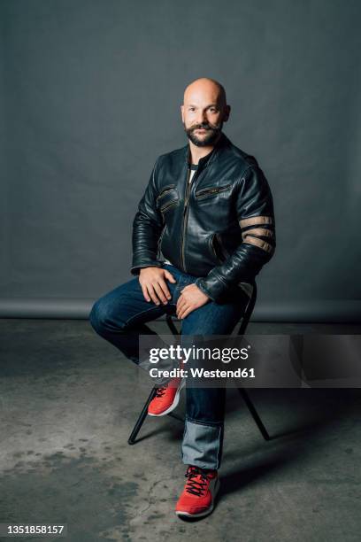 mid adult man sitting on chair in front of gray backdrop - chaqueta de piel imágenes fotografías e imágenes de stock