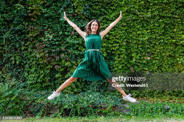 playful woman with arms outstretched jumping in front of ivy plants at park - frau springt hüpft stock-fotos und bilder