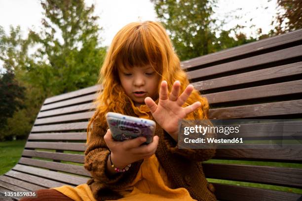 redhead girl using mobile phone while sitting on park bench - down syndrome girl stock pictures, royalty-free photos & images