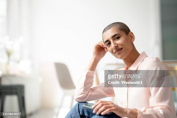 portrait of woman sitting on the floor in a loft holding smartphone - shaved head stock pictures, royalty-free photos & images