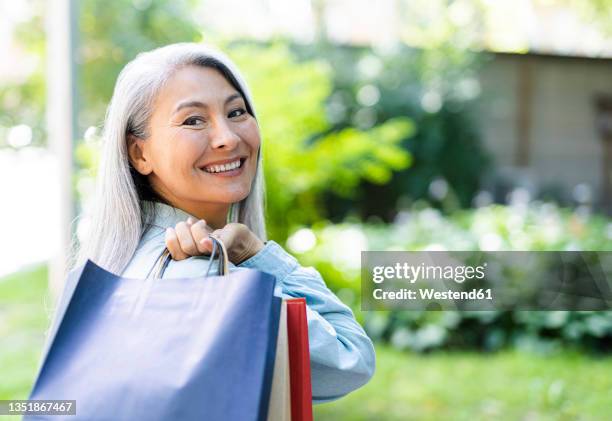 happy mature woman looking over shoulder while carrying shopping bags - one grey hair stock pictures, royalty-free photos & images