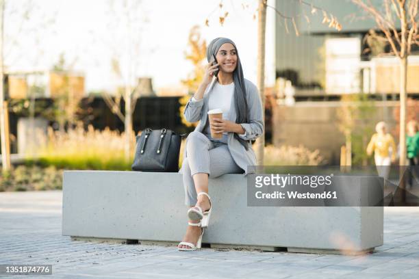 young businesswoman talking on smart phone while sitting on bench - personas en el fondo fotografías e imágenes de stock