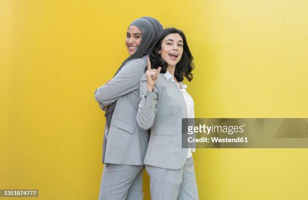 smiling female colleagues standing back to back by yellow wall - rücken an rücken stock-fotos und bilder