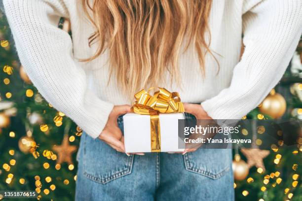 woman hiding a small white gift box behind her back in front of illuminated christmas tree - secret santa stock-fotos und bilder