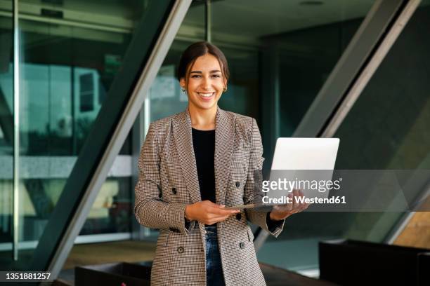 smiling businesswoman holding laptop out of office - standing with laptop imagens e fotografias de stock