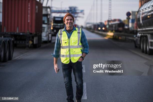 smiling male truck driver with hand in pocket walking on street at dock - truck driver stock pictures, royalty-free photos & images