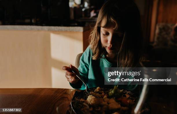 a young girl sits at a kitchen table and eats her dinner. scene is tranquil and calm. - picky eater stock pictures, royalty-free photos & images