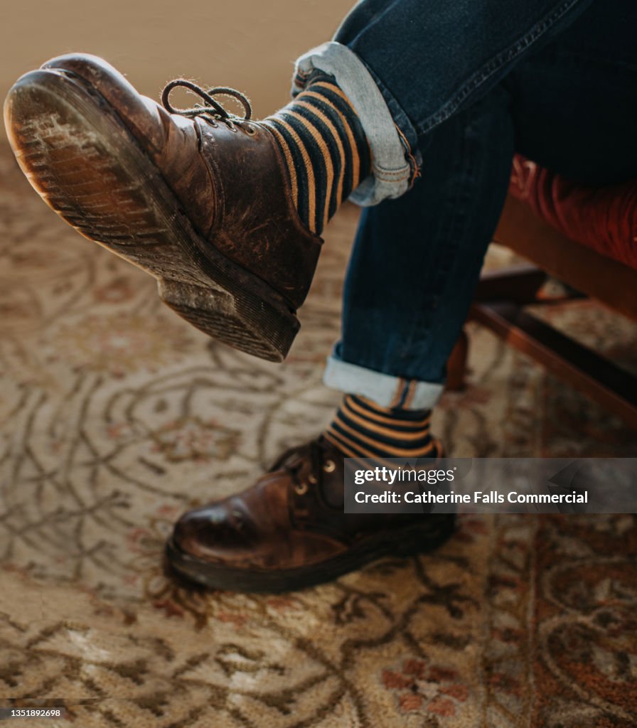 A man sits cross legged with focus on his classic style leather brown round toed shoes, and striped socks.