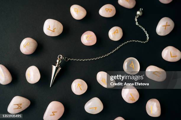 silver dowsing pendulum and many runes made of rose quartz with gold text arranged on black background. concept of magic esoteric rituals. macro photography in flat lay style - pendel stock-fotos und bilder