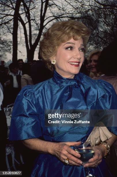 American actress Anne Baxter , wearing a blue dress, and holding a wine glass at an event, circa 1980.