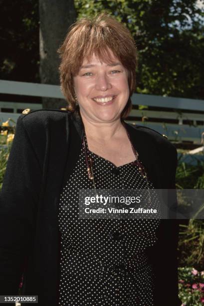 American actress Kathy Bates, wearing a a black jacket over a black outfit with white polka dots, a pair of glasses hanging from a cord around her...