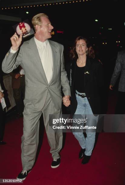 American actor Ed Begley Jr wearing a grey suit with a white shirt, holding hands with a woman as they attend the Westwood Premiere of 'Fatal...