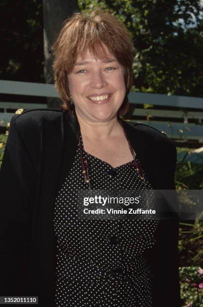 American actress Kathy Bates, wearing a a black jacket over a black outfit with white polka dots, a pair of glasses hanging from a cord around her...