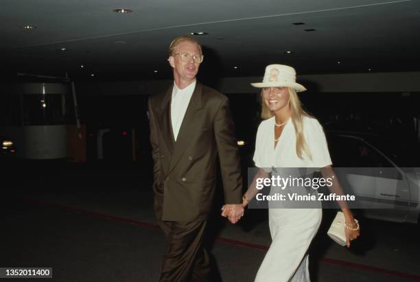 American actor Ed Begley Jr wearing a brown suit with a white shirt, holding hands with a woman who is dressed in white with a white hat, circa 1992.
