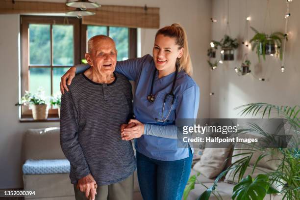 young female doctor walking with senior man indoors at home during home visit, looking at camera. - infirmière et maison photos et images de collection