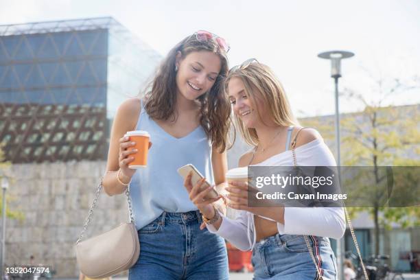 two young women with coffee cups watching on mobilphone in city - vest stock pictures, royalty-free photos & images